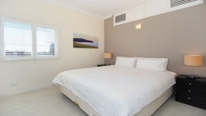 A bedroom with a large bed, two bedside tables with lamps, a window with blinds, and a painting on the wall. - Ultimate 2-Bed Apartment in Surry Hills - Ultimate Convenience (Surry Hills)