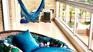 A wicker chair with a blue cushion and floral-patterned pillow, a blue hammock, and a view of greenery through large windows.