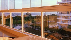 A modern room with large windows, a bed with a woven headboard, and a view of a cityscape and beach.