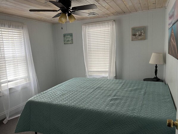 A bedroom with a bed, a ceiling fan, two windows with blinds, a small table with a lamp, and a wall decoration. - 3BR Beachfront Getaway – Behind Owner’s Home (Sylvan Beach)