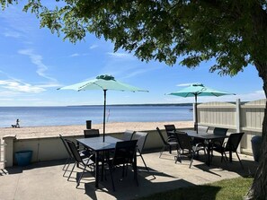 An outdoor patio with tables and chairs, two umbrellas, and a view of the beach and ocean. - 3BR Beachfront Getaway – Behind Owner’s Home (Sylvan Beach)