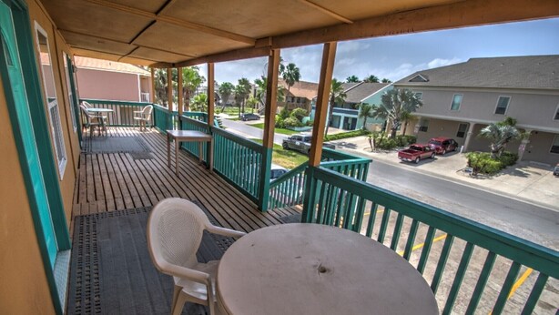 A covered outdoor area with a table and chairs, a view of a street with parked cars, and buildings in the background. - Oleander Beach Lodge - Four Condos for One Great Price (South Padre Island)
