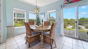A dining area with a round table, wicker chairs, and a view of a building through large windows. - Just Steps to The Beach, Surrounded by Nature – TAKING SNOWBIRD BOOKINGS NOW! (Orange Beach)