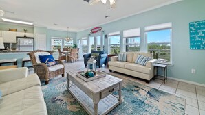 A spacious living room with a large window, a ceiling fan, and a kitchen area in the background. - Just Steps to The Beach, Surrounded by Nature – TAKING SNOWBIRD BOOKINGS NOW! (Orange Beach)