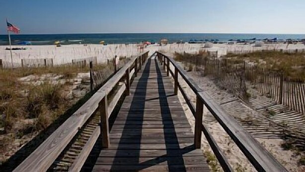 A wooden boardwalk leading to a beach with a sandy shore and an American flag. - Just Steps to The Beach, Surrounded by Nature – TAKING SNOWBIRD BOOKINGS NOW! (Orange Beach)