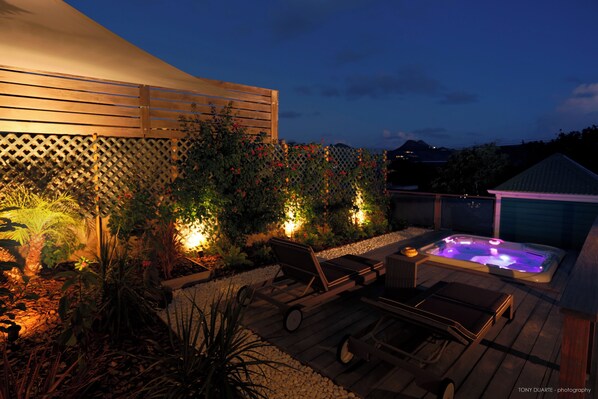 Une terrasse en bois avec un jacuzzi, des chaises longues et une pergola illuminée de guirlandes lumineuses.