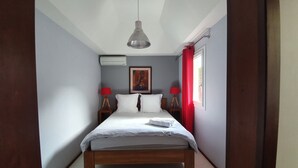 Un dormitorio con una cama, un lámpara colgante, dos mesitas de noche con lámparas, una ventana con cortinas y un equipo de aire acondicionado en la pared.