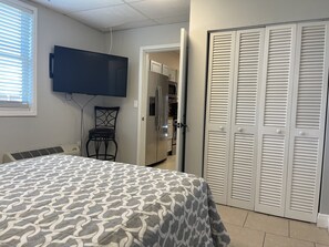 A bedroom with a bed, a TV mounted on the wall, a chair, a closet, and a refrigerator in the adjacent room. - NEW Apartment  Right Across the Street! With Parking Included! (Indialantic)