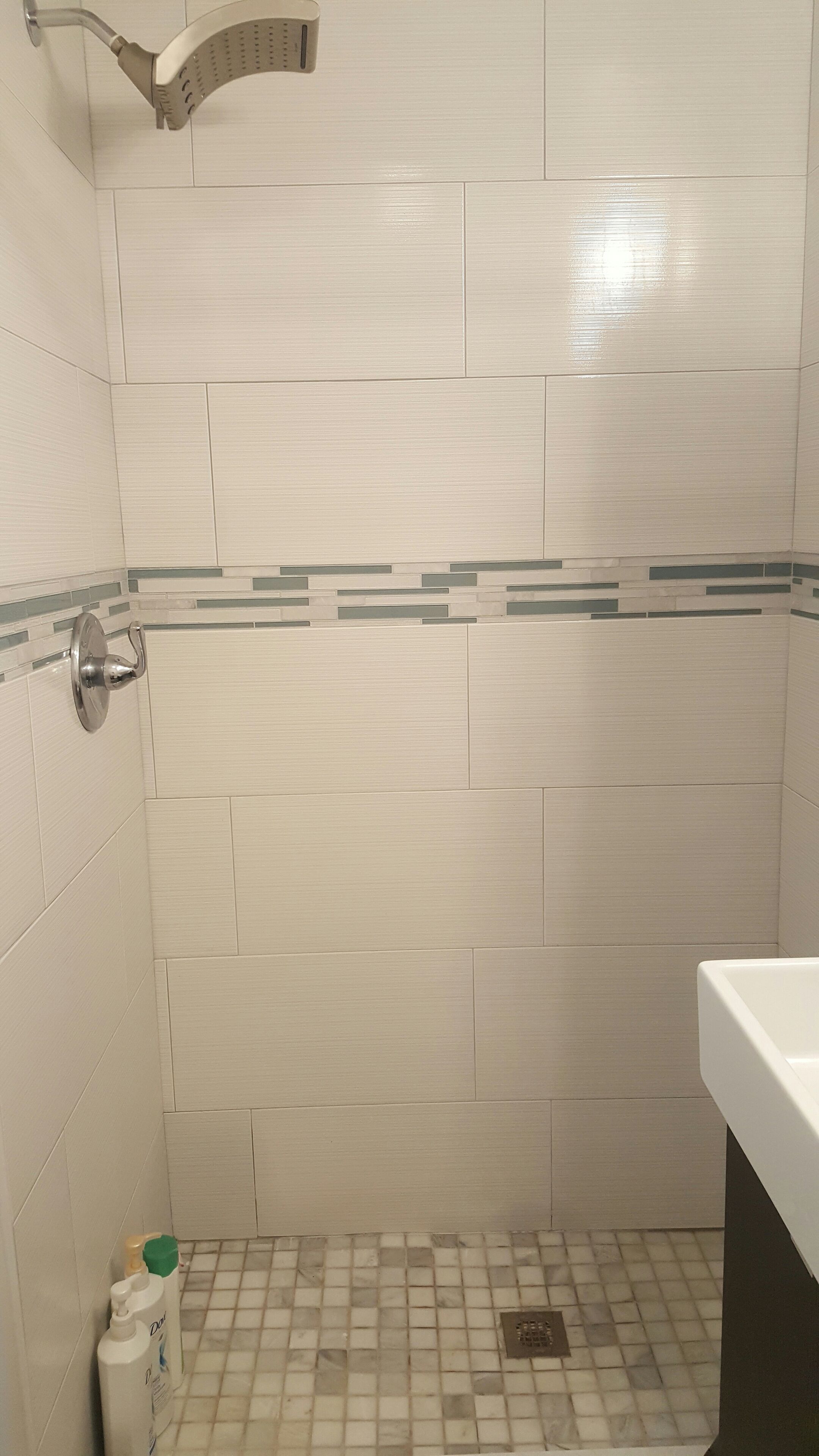 A bathroom with white tiles, a showerhead, a toilet, and a small bottle of lotion on the floor.