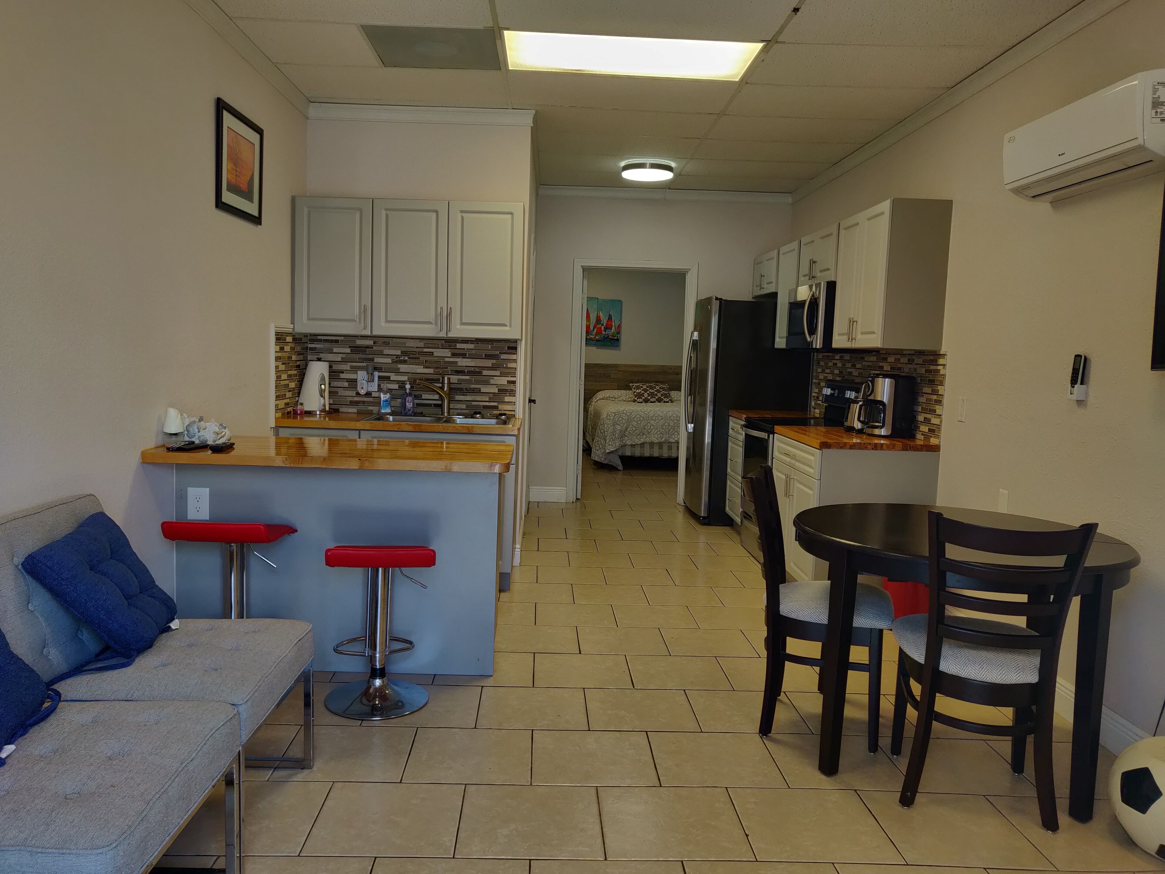 Cocina compacta con una isla central, comedor con mesa y sillas, sofá con un taburete rojo y una cama al fondo.