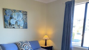 Un dormitorio con cama, mesita de noche, lámpara, ventana con cortinas y un cuadro en la pared.