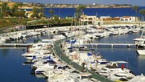A marina with numerous boats docked, a clear view of the sea, and a coastal landscape with buildings and palm trees in the background.