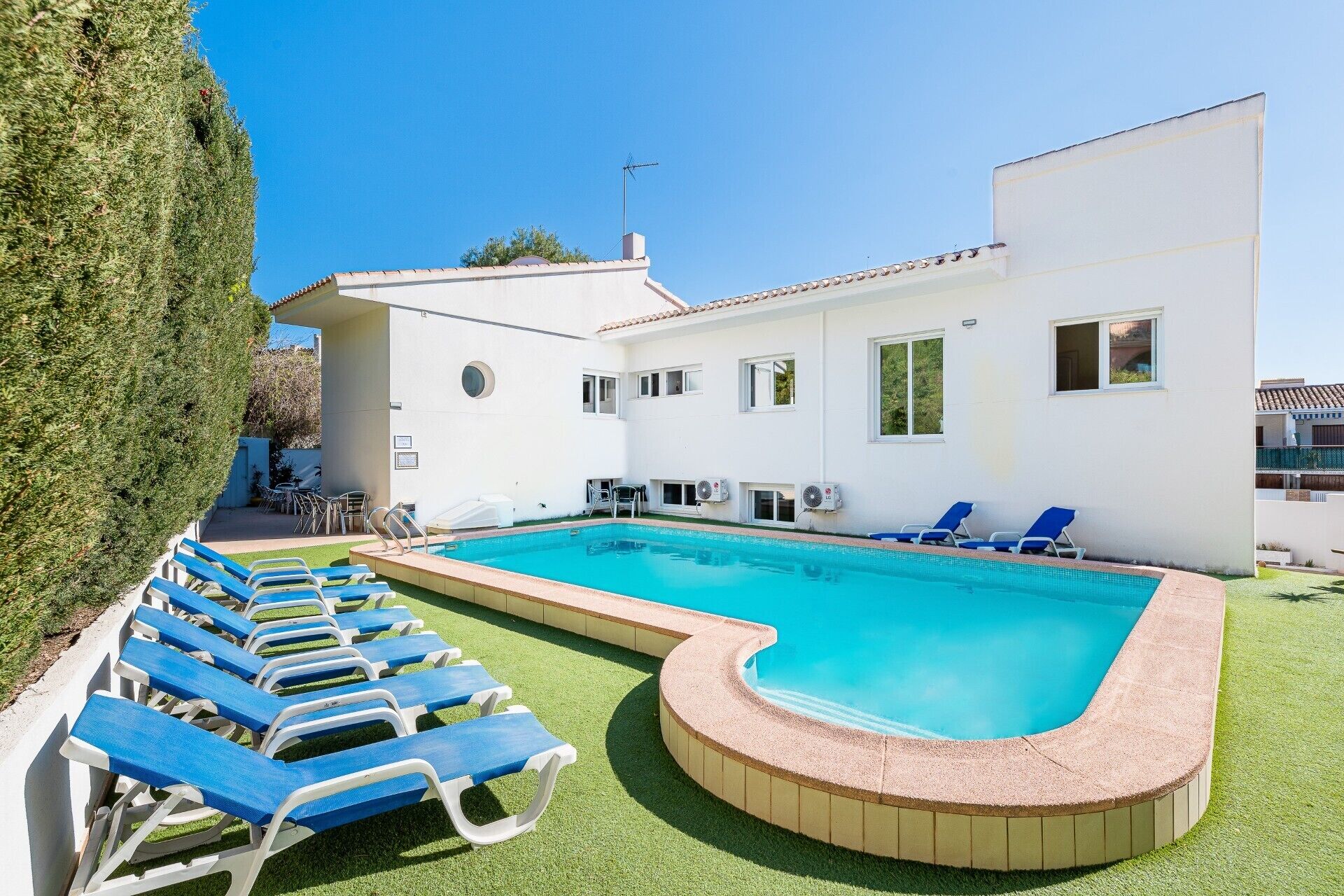 A modern pool area with a rectangular pool, blue lounge chairs, and a white building with multiple windows and air conditioning units.