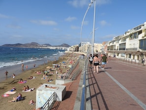 Una playa con gente tomando el sol y nadando, un paseo marítimo con barandilla de metal y edificios al fondo.