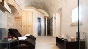 Un cuarto de spa con una silla de masaje, una mesa de vidrio con un frutero, una mesita auxiliar de mimbre negro, un box de ducha de vidrio y una puerta de madera.
