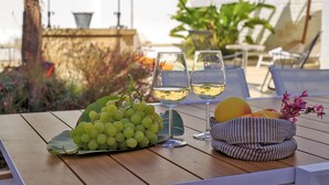 Una mesa con un racimo de uvas verdes, dos copas de vino blanco y un bol con frutas y flores.