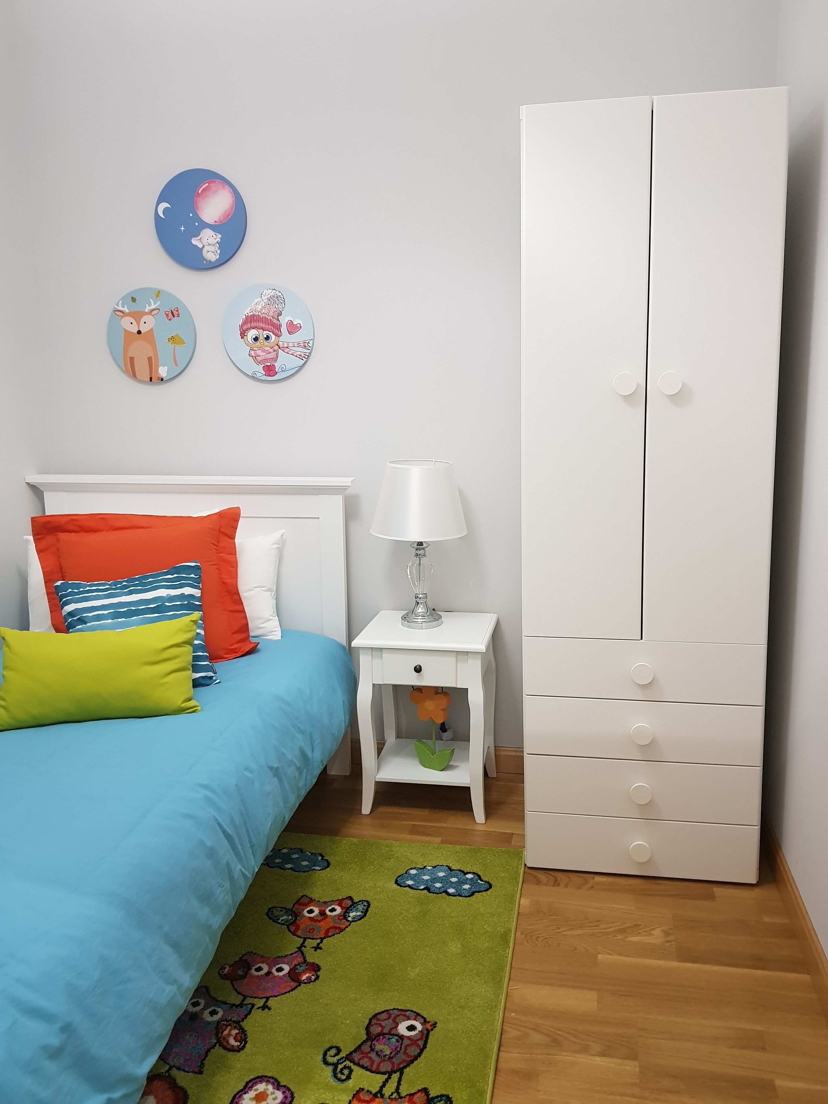 A bedroom with a bed, pillows, a nightstand, a lamp, a wardrobe, and wall decorations.