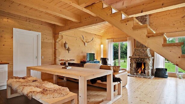 Ein gemütliches Blockhaus-Interieur mit Kamin aus Stein, Holzmöbeln und einem großen Fenster mit Blick auf einen Garten.