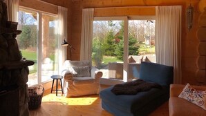 Ein gemütliches Blockhaus-Interieur mit Holzbalkendecke, Kamin aus Stein, einem blauen Sofa, einem weißen Sessel und einem großen Fenster mit Blick auf ein bewaldetes Gebiet.