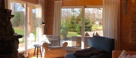 L’intérieur chaleureux d’un chalet avec un plafond en bois, une cheminée en pierre, un canapé bleu, un fauteuil blanc et une grande fenêtre donnant sur une zone boisée.
