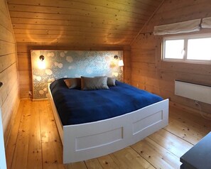 Et soveværelse med træloft, en seng med blåt sengetæppe, to vægmonterede lamper og et vindue med hvidt gardin.