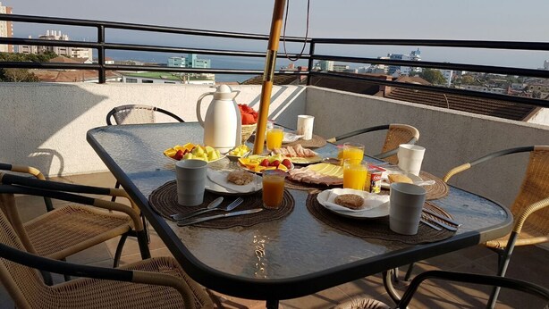 Mesa de desayuno en un balcón con sillas de mimbre, una sombrilla y vistas a la ciudad y al mar.