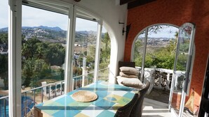 Un balcó amb vista a una vila i muntanyes, una taula amb un estriat i una cadira de vimetla.