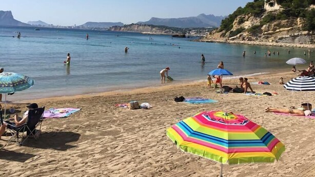 Una platja amb gent banyant-se i prenent el sol, sombrerets i un cel serè.