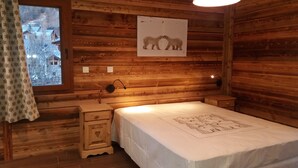 Ein Zimmer in einer Holzhütte mit einem Bett, einem Nachttisch, einer Wandlampe und einem Fenster mit Vorhang.