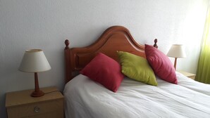 Uma cama bem arrumada com roupa de cama branca e duas almofadas, uma mesa de cabeceira de madeira com abajur e uma pequena cômoda.