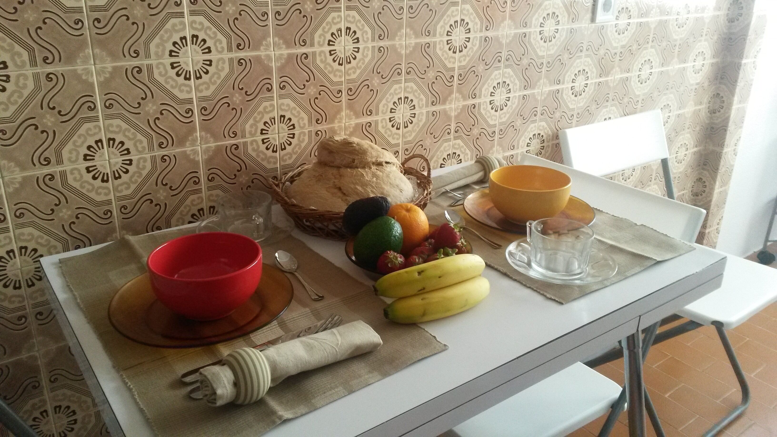 Uma bancada de cozinha com um pão, uma tigela de frutas, uma tigela vermelha e um copo de vidro.