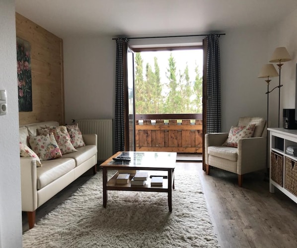 A cozy living room with a sofa, armchair, and coffee table, a wooden deck outside, and a view of trees.