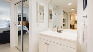 A modern bedroom with a large bed, a sliding door leading to a bathroom, and a large mirror above the sink. - Ocean Village Beach Condo, Golfcourse and Tennis Resort (Fort Pierce)