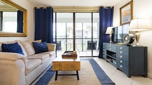 A living room with a sofa, coffee table, TV stand, and a view through a glass door. - Ocean Village Beach Condo, Golfcourse and Tennis Resort (Fort Pierce)