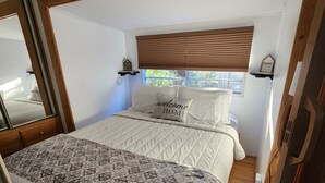 Een slaapkamer met een groot bed, houten hoofdeinde en een raam met zonwering. Er staan twee nachtkastjes met lampen en er hangt een spiegel aan de muur.