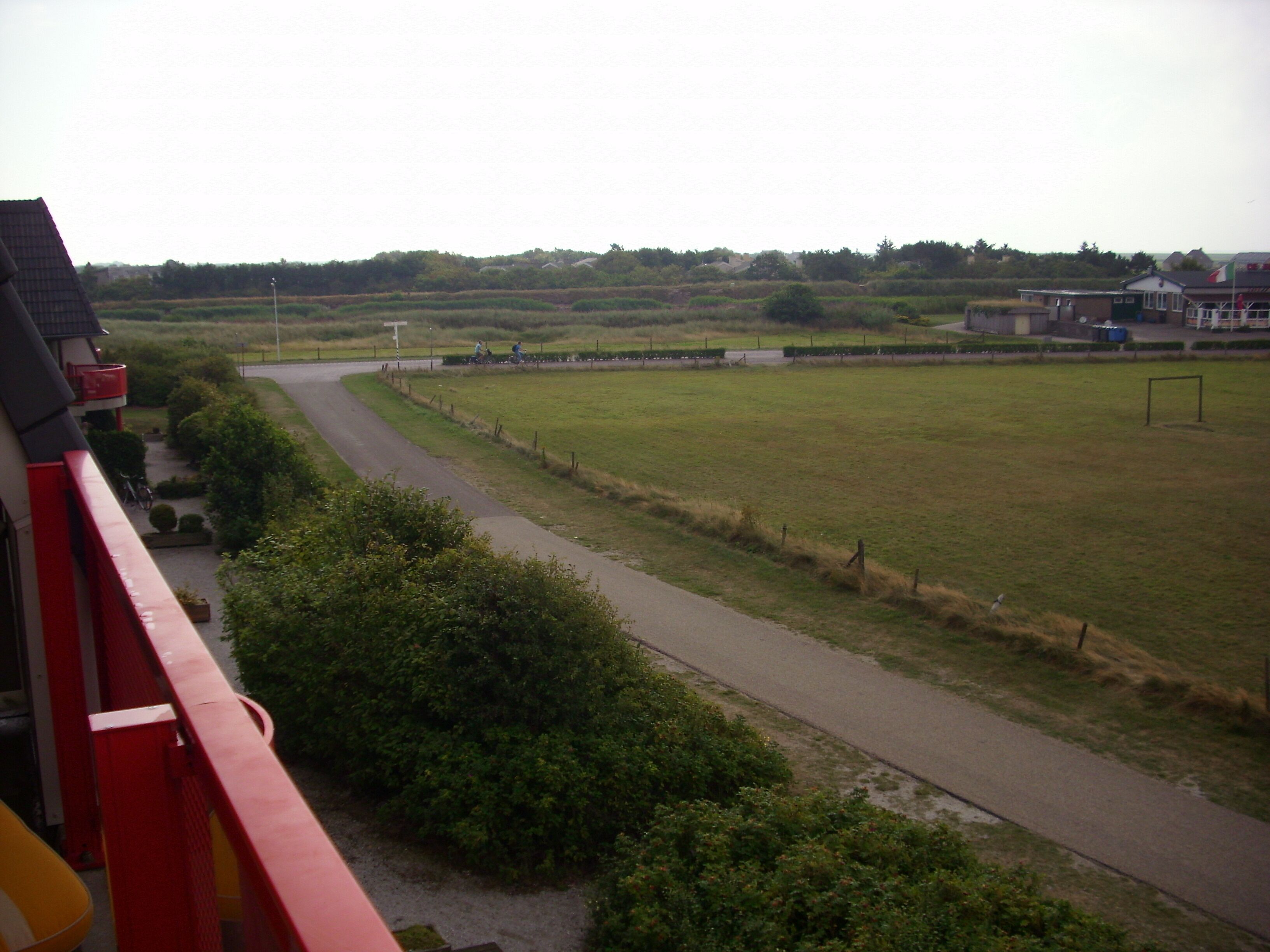 Une route de campagne sinueuse traverse un champ verdoyant, avec une clôture d’un côté et un bâtiment au toit rouge de l’autre.