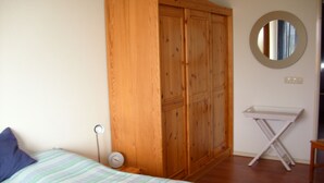 Un dormitorio con cama, un armario de madera, una mesita de noche con reloj y una pequeña estantería blanca.