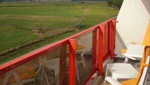 Un balcón con vista a un campo verde y una torre distante. Hay sillas blancas y una barandilla roja.