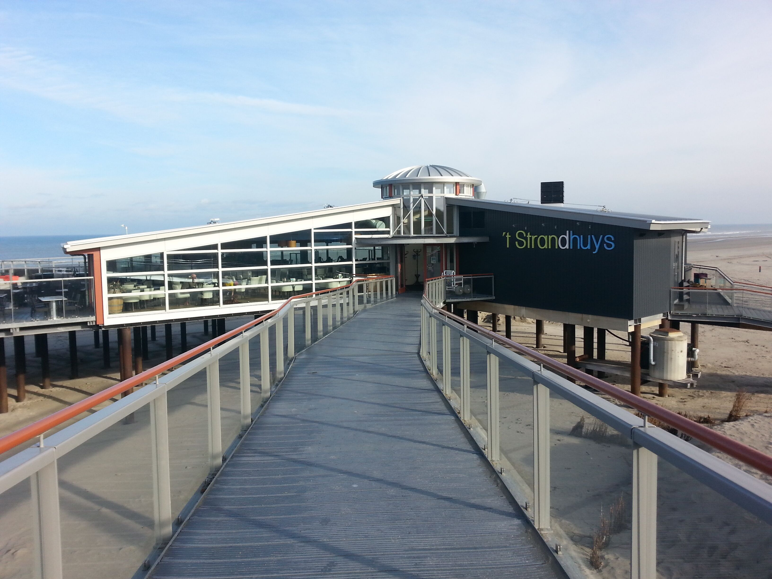 A boardwalk leading to a beachside building with a glass facade and a sign reading "Strandhuy's".