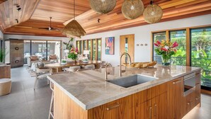 A modern kitchen with a large island, wooden cabinets, and a view of greenery outside. - Hale Luana By Parrish Kauai-beachfront estate with pool (Koloa)