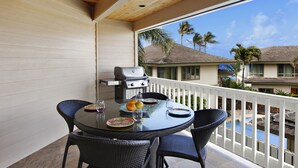 A balcony with a round table and chairs, a grill, and a view of palm trees and houses.