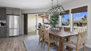 A modern kitchen with a stainless steel refrigerator, a wooden dining table with woven chairs, and a view of a balcony with a grill and plants.