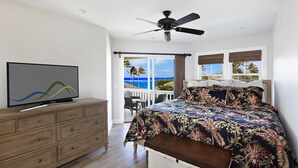 A bedroom with a bed, a TV, a ceiling fan, a wooden dresser, and a balcony with a view of the ocean.
