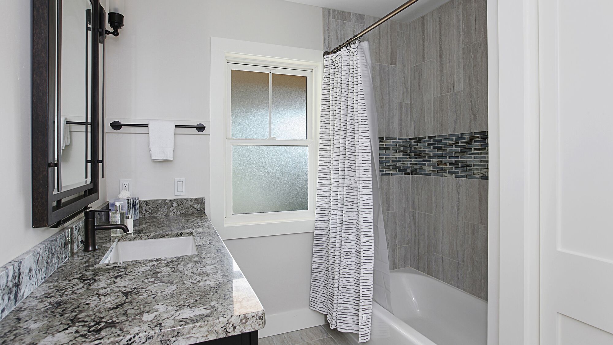 A bathroom with a granite countertop, a mirror, a towel rack, a frosted window, and a shower with a striped curtain.