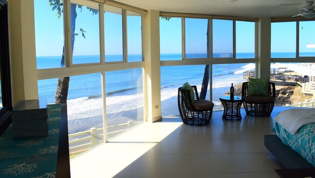 Une chambre avec de grandes fenêtres donnant sur la plage, un lit, une chaise et une petite table sur laquelle se trouvent une bouteille et un verre.