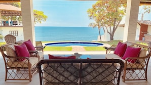 Un espace extérieur aménagé, abrité et doté de mobilier en osier et de coussins, offrant une vue sur une piscine et la mer.