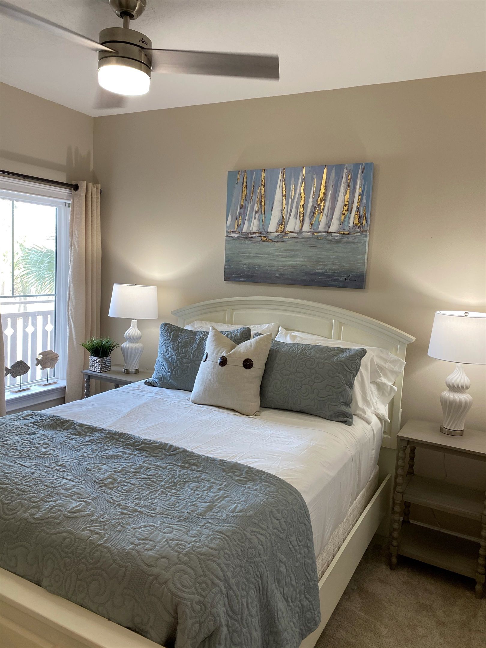 A neatly arranged bedroom with a bed, a ceiling fan, a painting of sailboats, and a window with curtains.