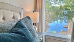 Un lit avec une tête de lit, une lampe et une vue sur une piscine et des palmiers, visible par une fenêtre.
