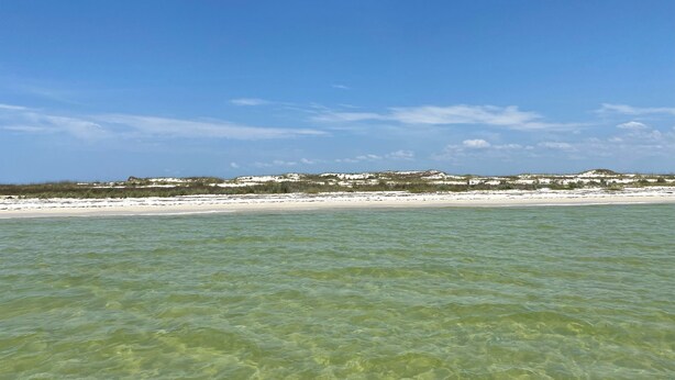 Une plage avec une eau claire et peu profonde, et une vue lointaine sur des dunes.