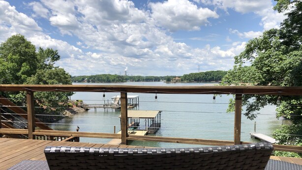 Une terrasse en bois, agrémentée de chaises en osier, offre une vue sur un lac paisible, des quais et une ligne électrique distante.